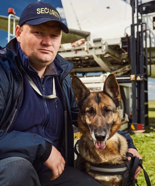 Security officer looking at camera with serious expression while wrapping arm around adorable German Shepherd dog